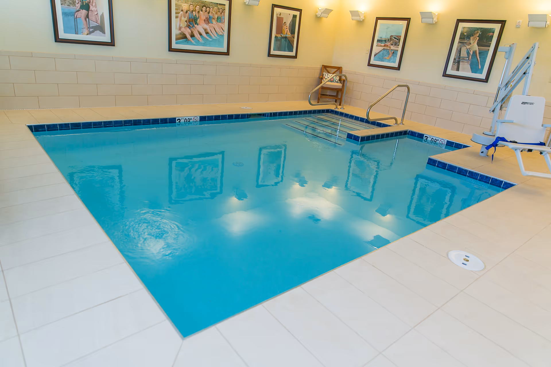 Indoor swimming pool with clear blue water, surrounded by beige tiled flooring and walls. The pool has metal handrails and steps for entry. Several framed pictures of swimmers are hung on the walls. A pool lift chair is positioned on the right side for accessibility.
