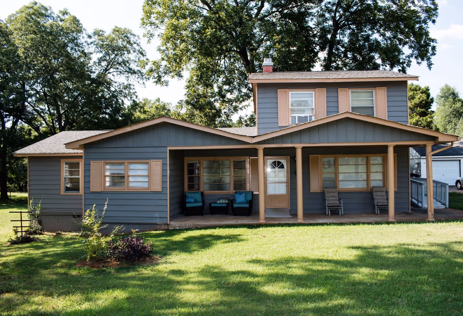 Front exterior view of a two-story house painted in blue with beige trim and shutters. The house features a covered porch with seating including two chairs with blue cushions and two wooden chairs. The lawn is green and well-maintained with some small plants and trees around the property.