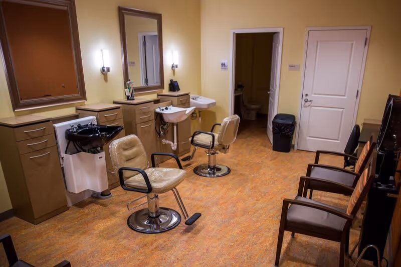 Interior view of a salon area in a senior living facility with two salon chairs in front of sinks and mirrors, additional seating with chairs along the right wall, and a doorway leading to a restroom.