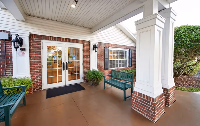Covered entrance area of a building with double glass doors, brick walls, white columns, two green benches, potted plants, and outdoor lighting fixtures.