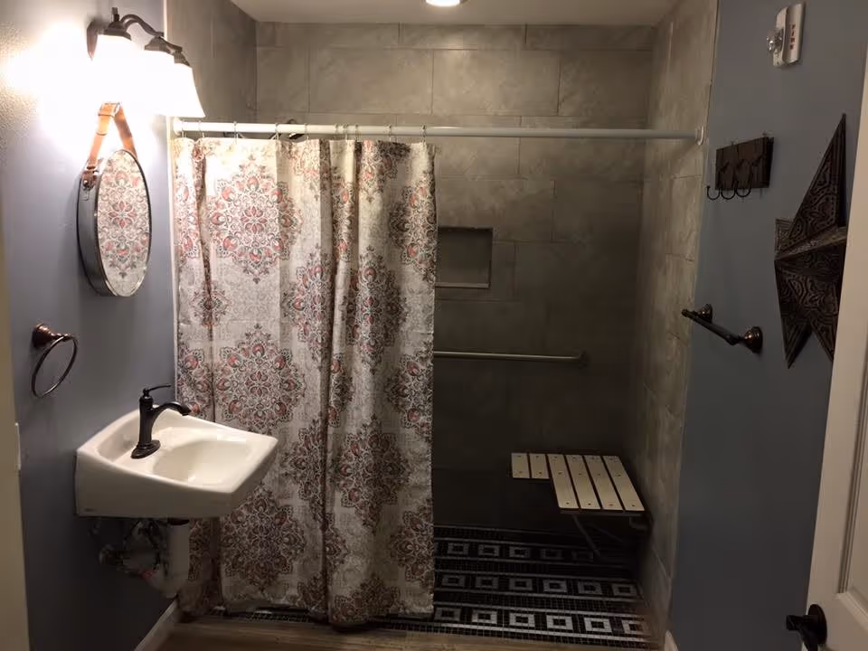 A bathroom with a small white sink and black faucet on the left wall, a decorative round mirror above the sink, and a patterned shower curtain covering a walk-in shower area with tiled walls and a foldable shower bench. The floor has a patterned tile design, and there are towel holders and a decorative star on the right wall.