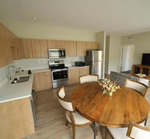Open-plan kitchen and dining area with light wood cabinets, stainless-steel appliances, and a round wooden table with chairs.
