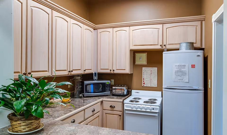 A compact kitchen with light wood cabinets, a white electric stove, a white refrigerator with a notice taped on the door, a microwave, a toaster, and a potted green plant on the countertop.