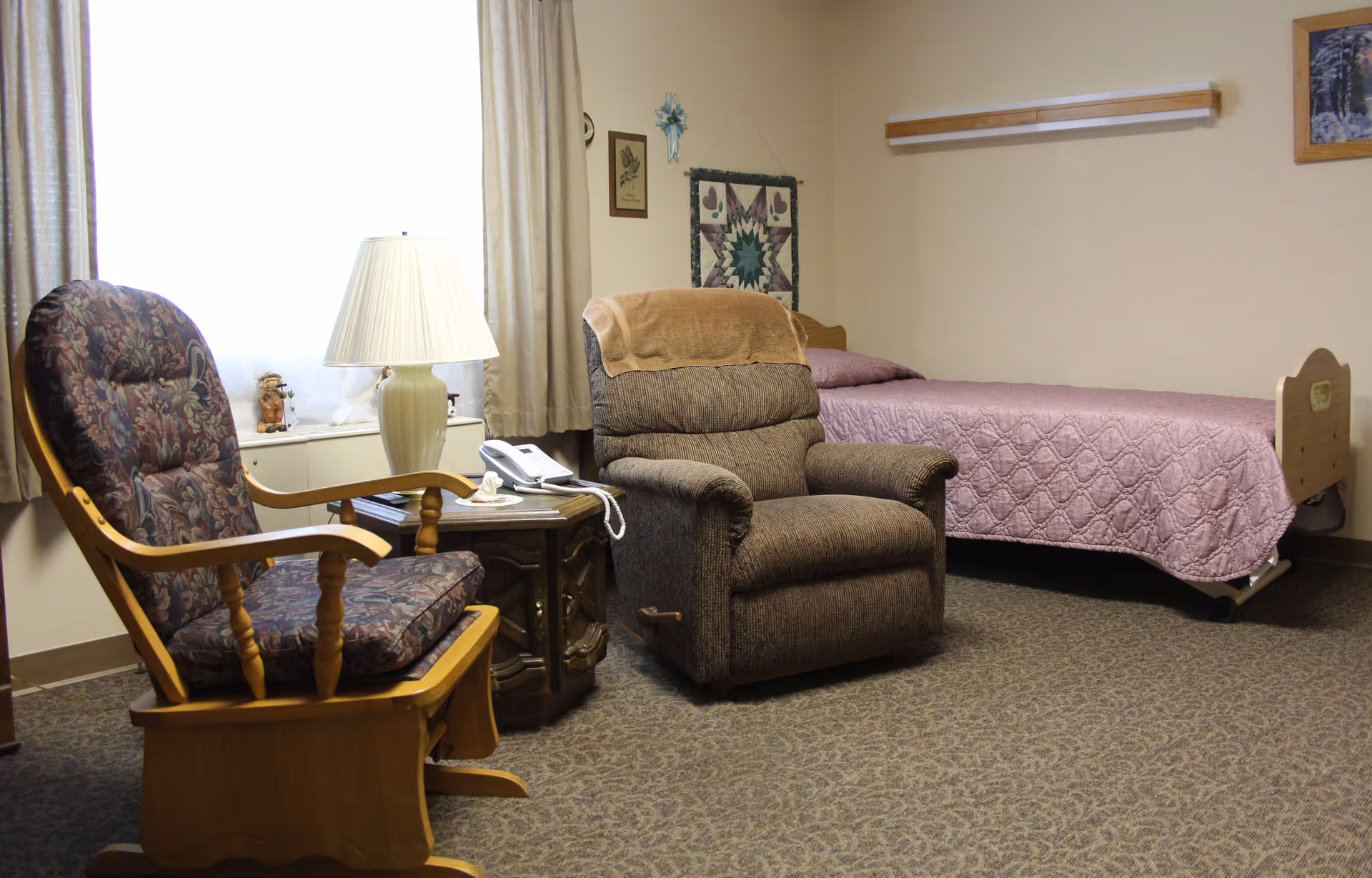 A cozy senior living bedroom featuring a single bed with a pink quilt, a brown recliner chair with a tan blanket draped over the back, a wooden rocking chair with floral cushions, a small wooden side table with a white lamp and a telephone, beige walls decorated with framed artwork and a quilt, and a window with beige curtains letting in natural light.