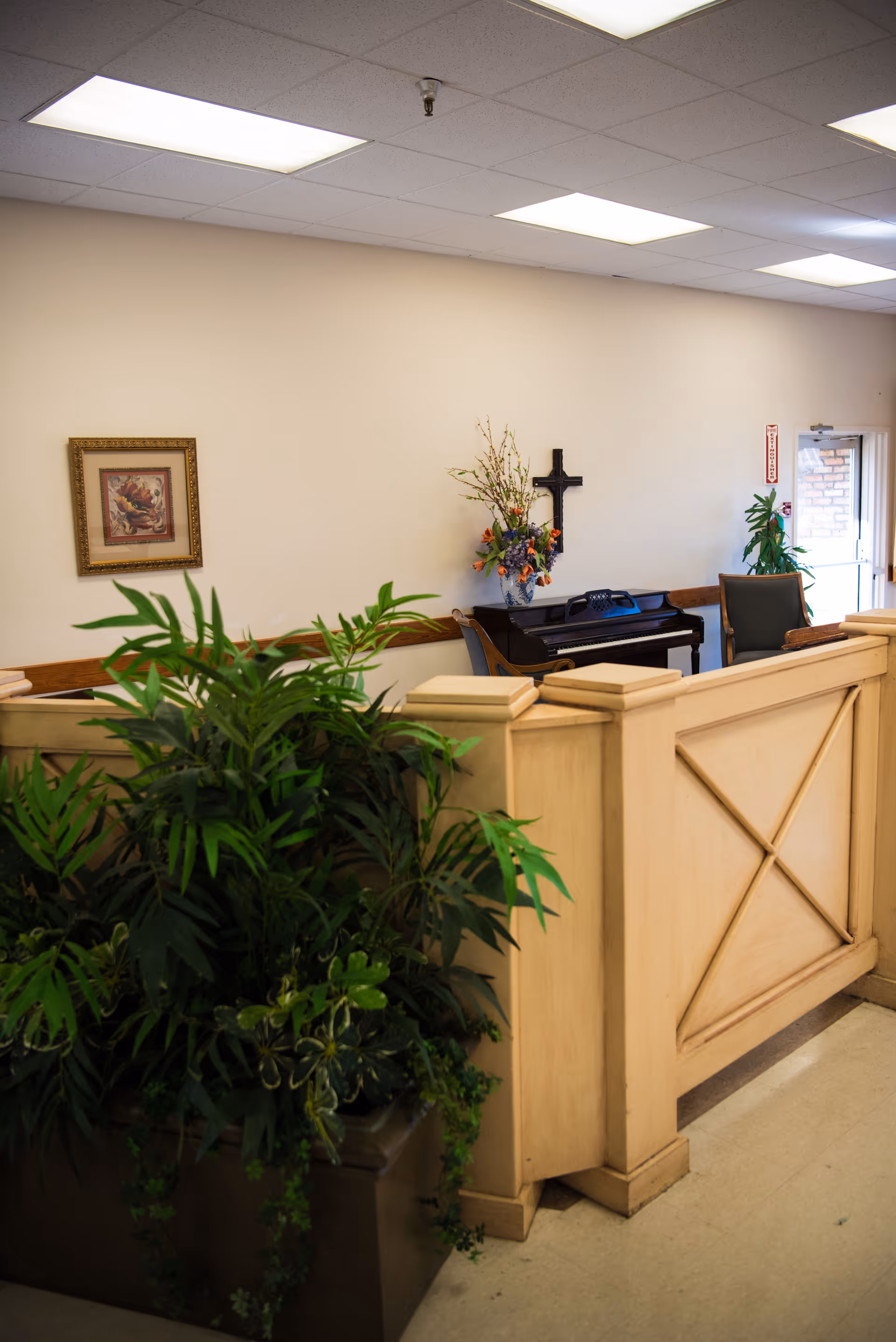 Bright interior reception area with a wooden partition, large potted plants, a piano, and wall decorations including a cross and framed art.