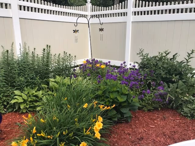 A garden area with various green plants and flowers including yellow and purple blooms, surrounded by a white fence with a picket top. There is a metal garden hook with two hanging wind chimes shaped like dragonflies.