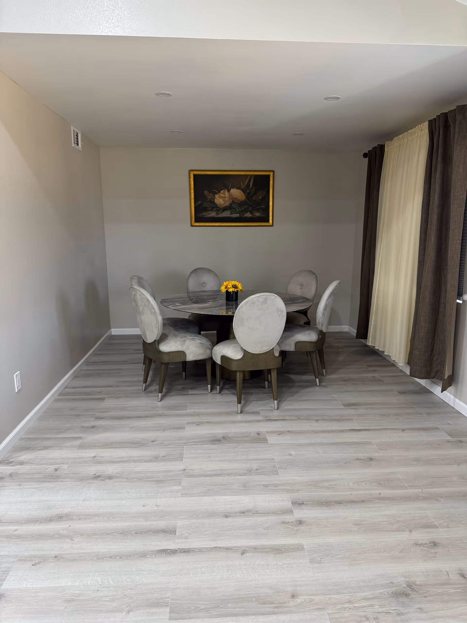 Dining area with a round table surrounded by six upholstered chairs, a small vase of yellow flowers, a framed painting on the wall, and curtains at right.