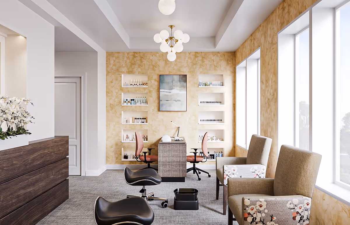 A bright and modern salon area in a senior living facility with two pink swivel chairs facing a small table with a lamp and nail care tools. The back wall has built-in shelves displaying various beauty products and a framed abstract painting. Two beige armchairs with floral patterned armrests are positioned near large windows letting in natural light. The floor is carpeted and the ceiling features a decorative light fixture.