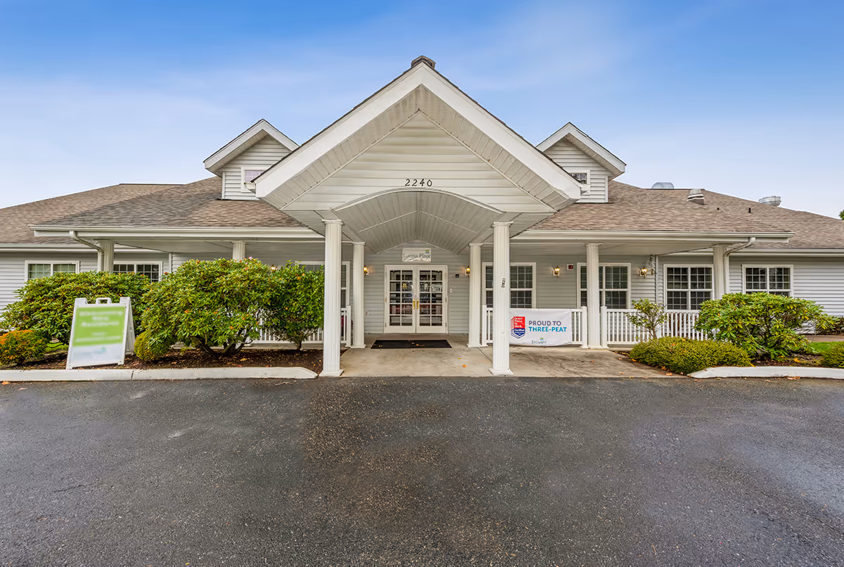 Front exterior view of a single-story senior living facility building with a covered entrance supported by white columns. The building is light gray with white trim and has bushes and greenery on either side of the entrance. There are signs near the entrance, including one that reads 'Proud to Three-Peat'. The sky is clear and blue.