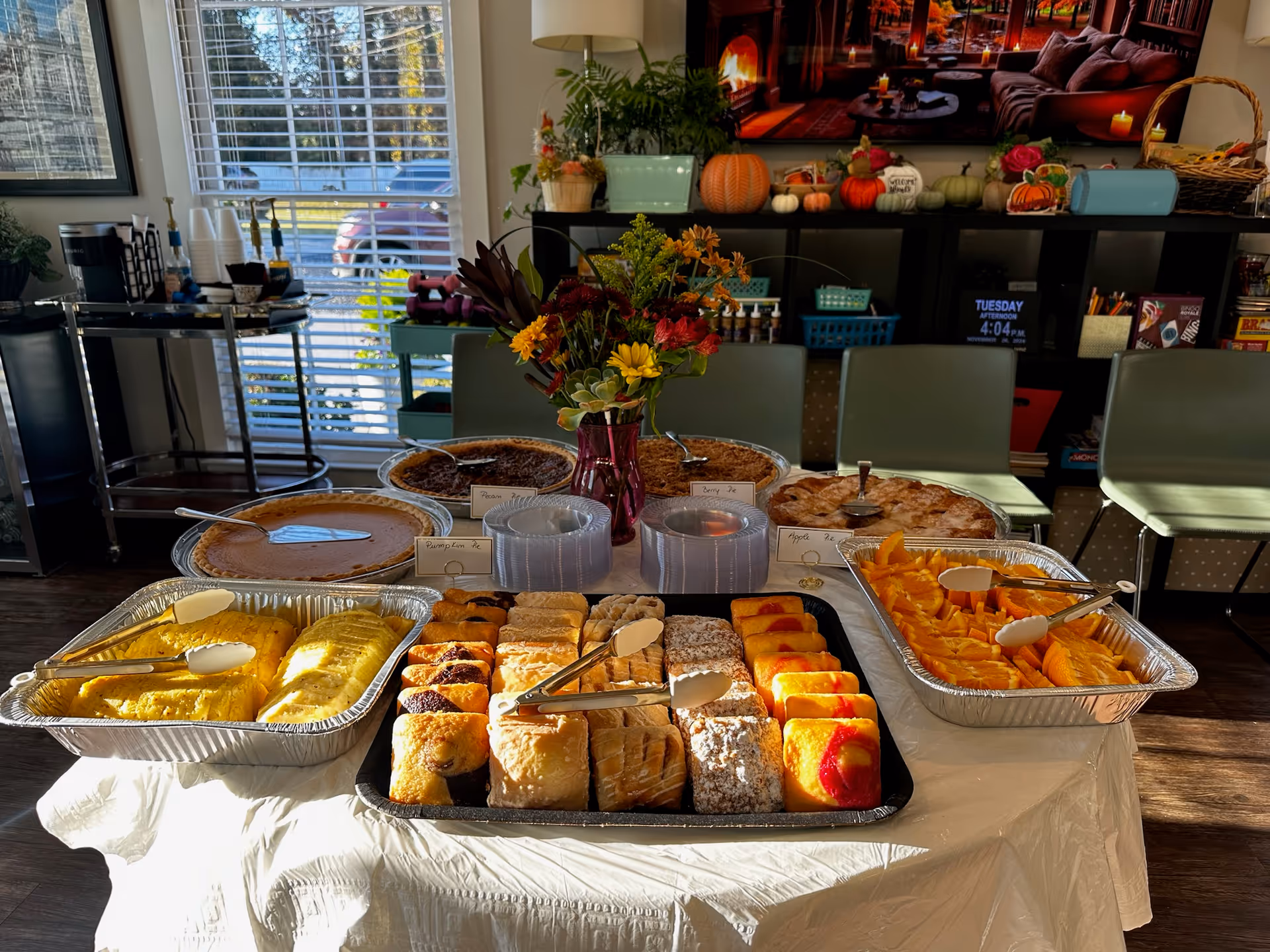 Buffet table in a communal dining area displaying assorted pastries, pies, sliced fruit and a vase of flowers.