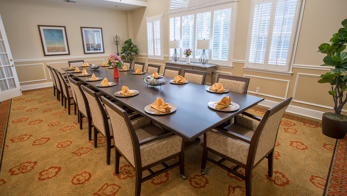 A formal dining room with a long dark wooden table set for twelve people. Each place setting has a plate with a folded beige napkin. The room has beige walls with white trim, two framed pictures on one wall, large windows with white blinds, a sideboard with two lamps and a flower vase, and a patterned carpet with red and beige tones. There is a potted plant in the corner.
