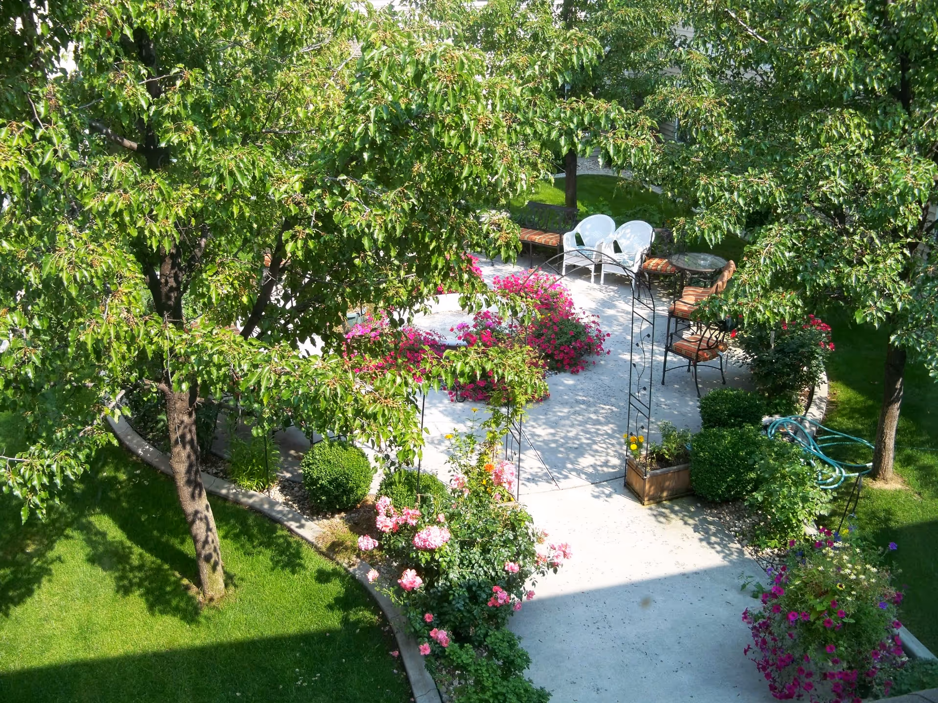 A sunny outdoor garden area with a concrete patio surrounded by green trees, bushes, and colorful flowers. The patio has several chairs and a small table arranged for seating. A garden hose is coiled on the grass nearby.