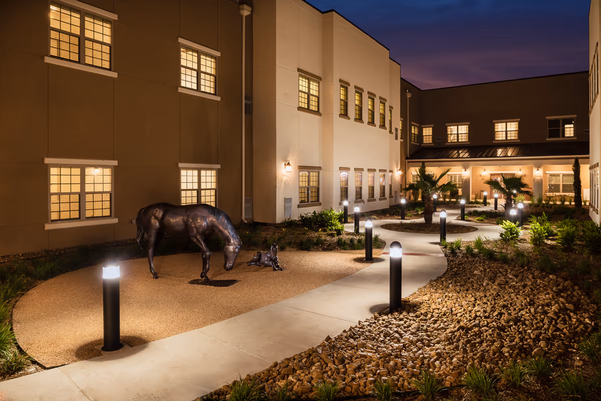 Night view of an outdoor courtyard at a senior living facility with illuminated pathway lights, landscaped plants, and sculptures of a horse and a foal. The building surrounding the courtyard has multiple windows with warm lights inside.