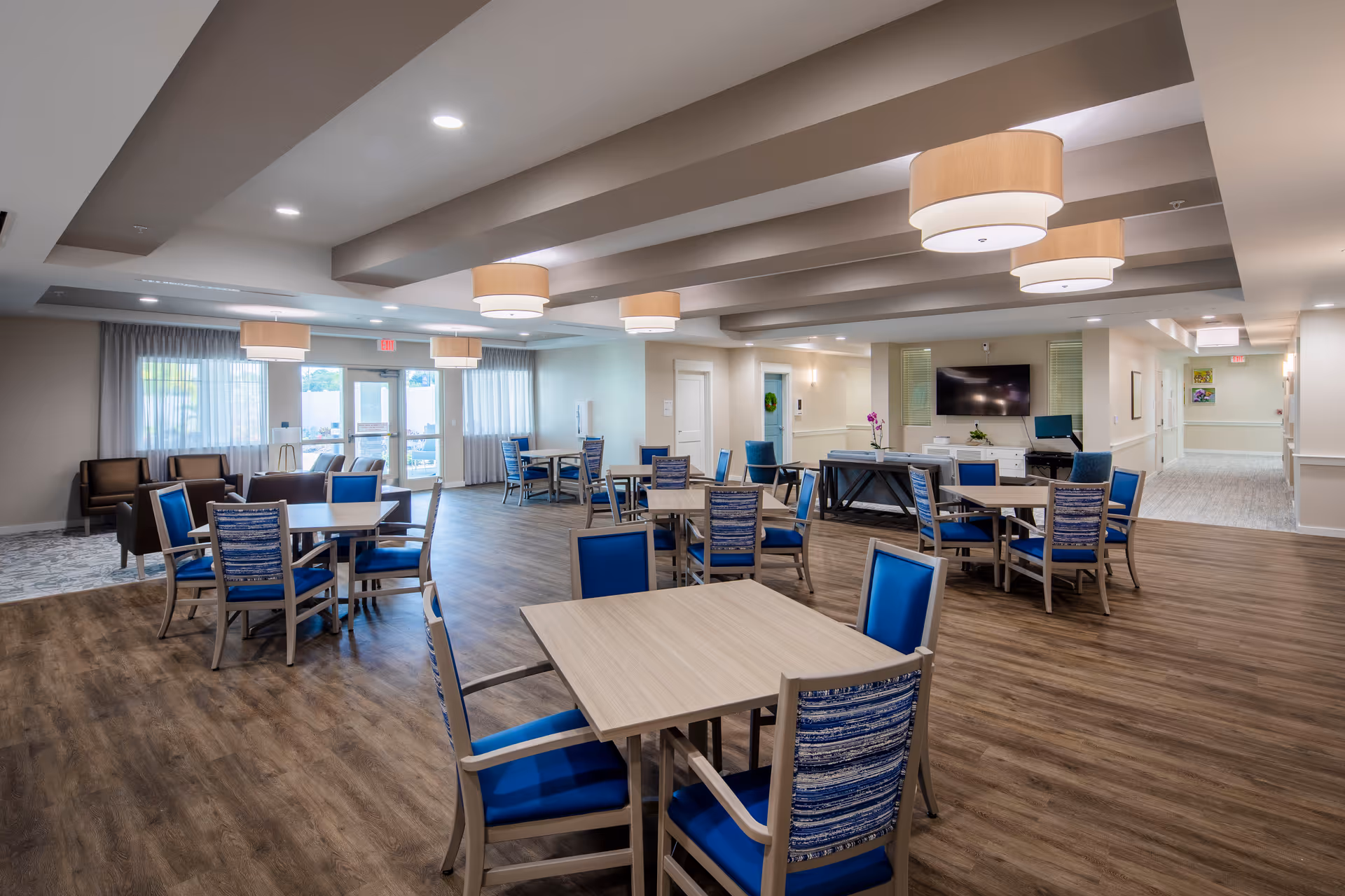 Spacious communal dining and activity room with multiple tables and blue-upholstered chairs, wood-look flooring, pendant lights, and a wall-mounted TV.