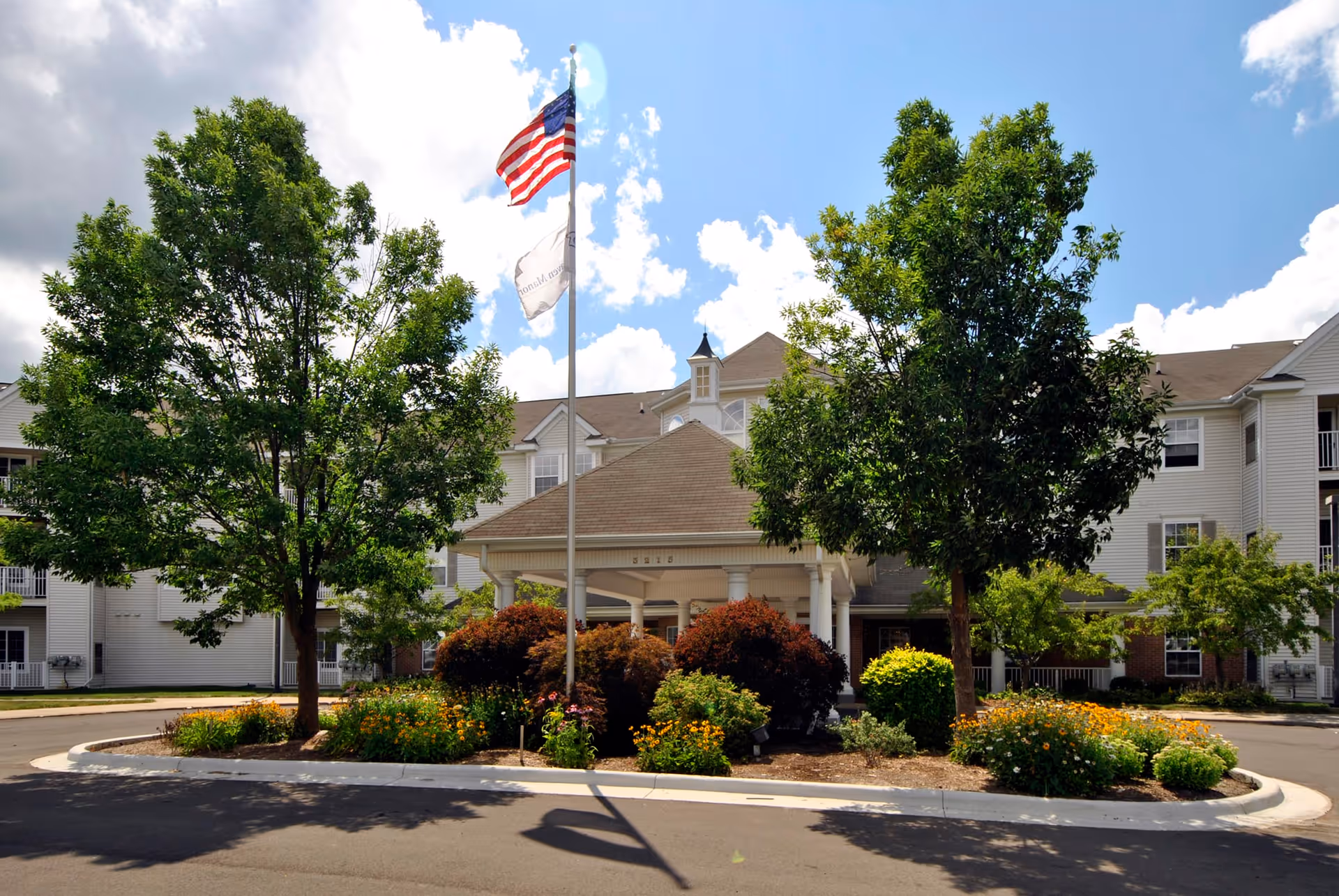 Front exterior view of a senior living facility named Grandhaven Manor with a covered entrance, two flagpoles displaying the American flag and another flag, surrounded by trees and landscaped bushes and flowers under a partly cloudy sky.