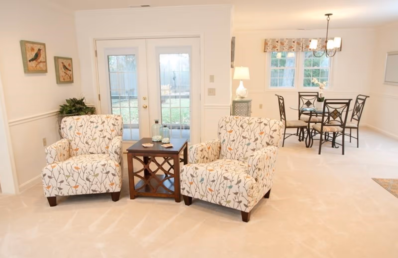 A bright living area with two patterned armchairs featuring a floral and bird design, separated by a wooden side table with decorative glass bottles. Behind the chairs, there are double glass doors leading outside. To the right, there is a dining area with a round glass table and four metal chairs with cushioned seats, under a hanging light fixture. The walls are light-colored with two framed bird pictures on the left wall and a lamp on a small table near the dining area.