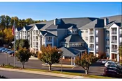 Front exterior of a multi-story retirement community building with a covered entrance, parked cars, and trees lining the driveway.