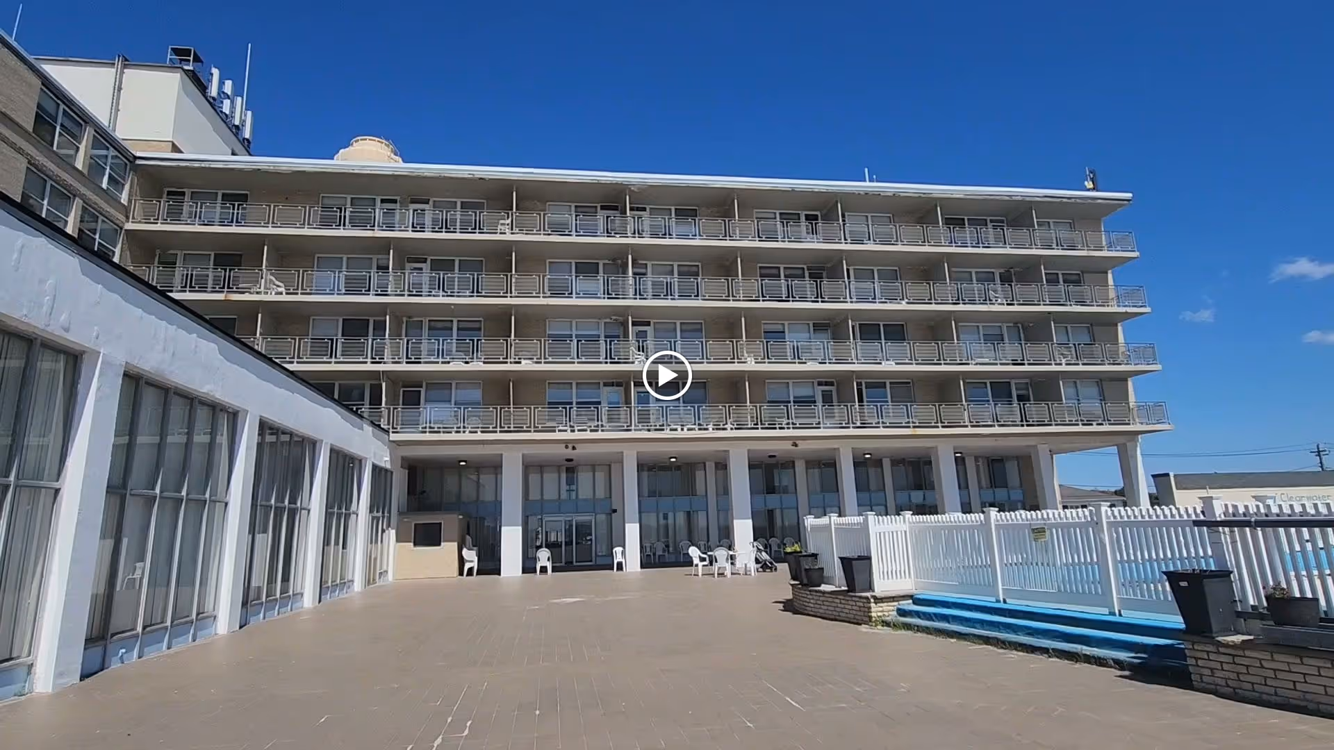 Exterior view of a multi-story building with balconies on each floor under a clear blue sky. The building has a large open patio area in front with white chairs and a white fence on the right side.