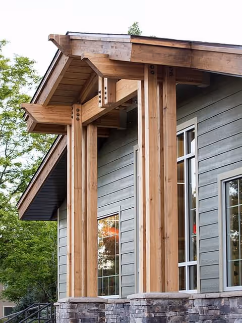 Close-up view of the exterior corner of a building featuring large wooden beams supporting an overhanging roof, with gray horizontal siding and stonework at the base. Trees and greenery are visible in the background.