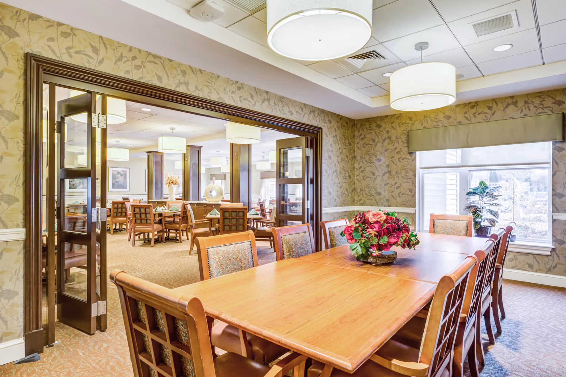 A well-lit dining room in a senior living facility featuring a long wooden table with cushioned chairs around it. A floral centerpiece decorates the table. The room has patterned wallpaper, a large window with a green valance, and ceiling lights. Through an open doorway, another dining area with multiple tables and chairs is visible.