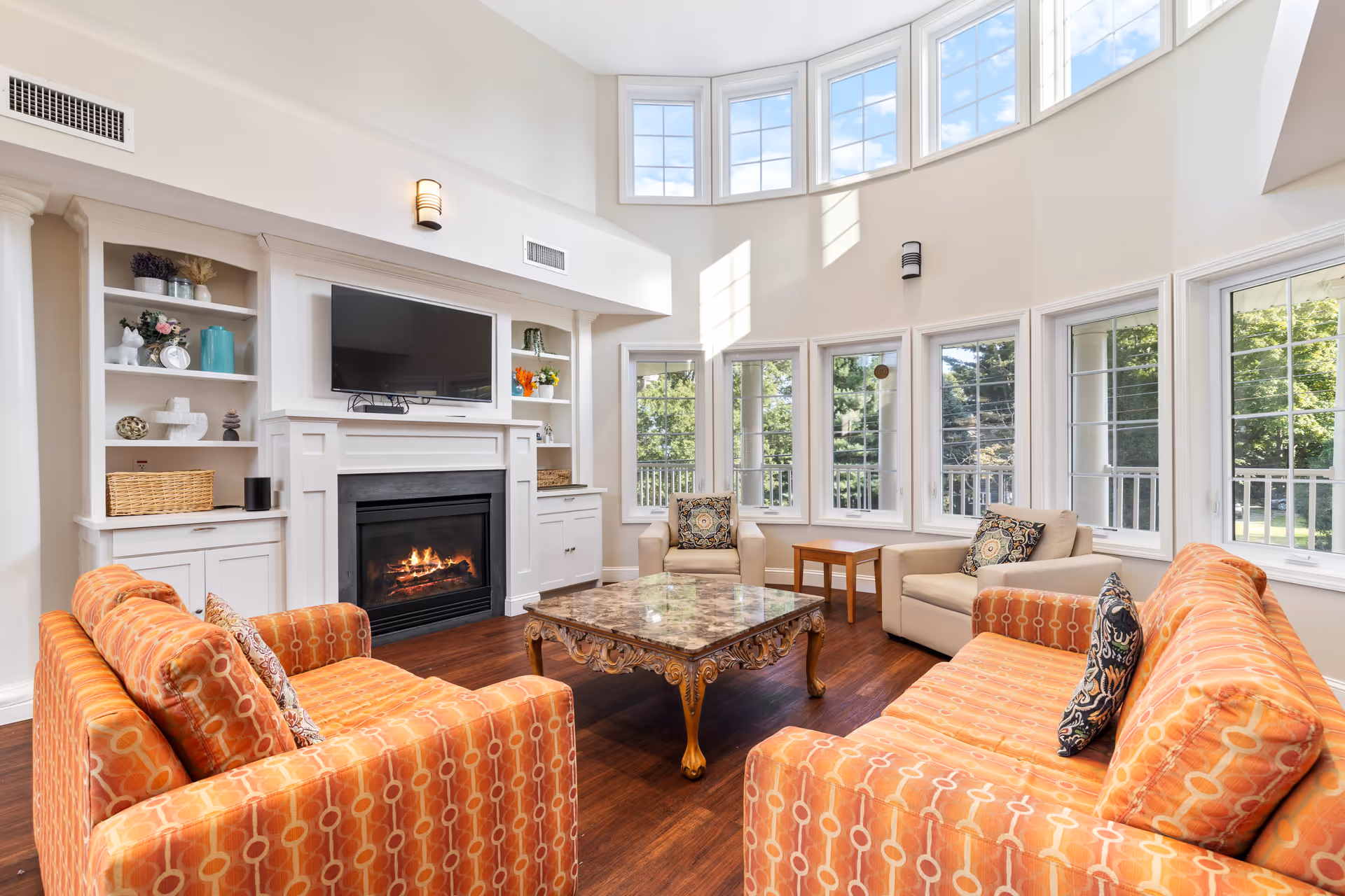 A bright and spacious living room with high ceilings and large windows letting in natural light. The room features two orange patterned sofas, two beige armchairs with decorative pillows, a marble-top coffee table with ornate wooden legs, a fireplace with a mounted flat-screen TV above it, and built-in white shelves with decorative items. Outside the windows, greenery and trees are visible.
