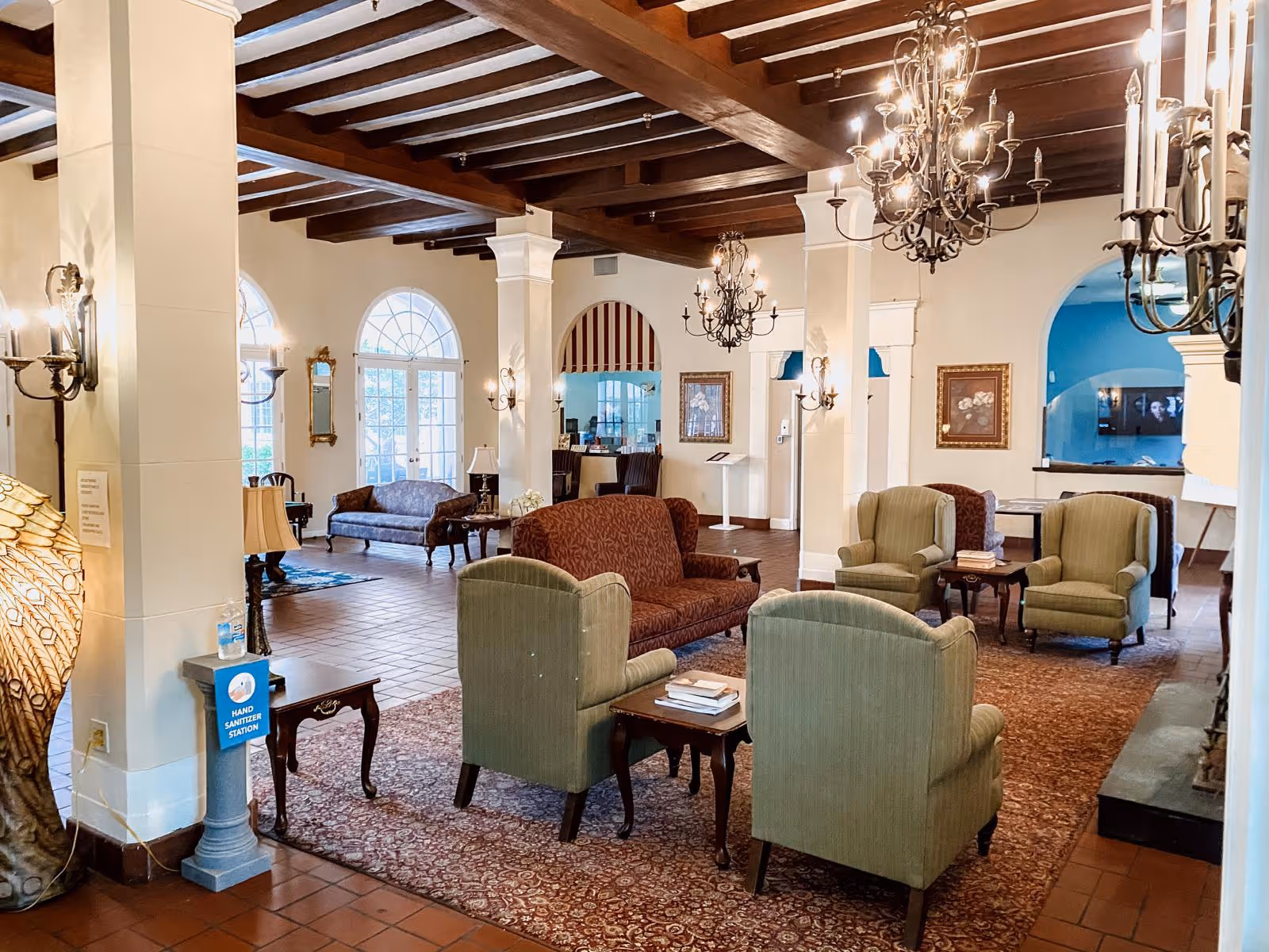 A spacious and elegant common area with vintage-style furniture including green armchairs and a red patterned loveseat arranged around small wooden tables on a large area rug. The room features exposed wooden ceiling beams, multiple chandeliers, wall sconces, large arched windows and doorways, and a hand sanitizer station near a pillar.