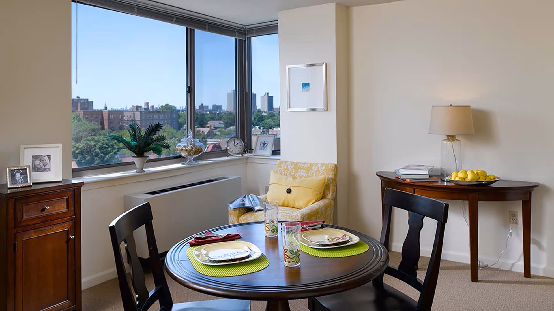 A cozy dining area with a round wooden table set for two with plates, glasses, and napkins. Two dark wooden chairs are positioned around the table. In the background, there is a yellow patterned armchair with a cushion, a wooden side table with a lamp, books, and a bowl of lemons. Large windows provide a view of a cityscape with buildings and trees under a clear blue sky.