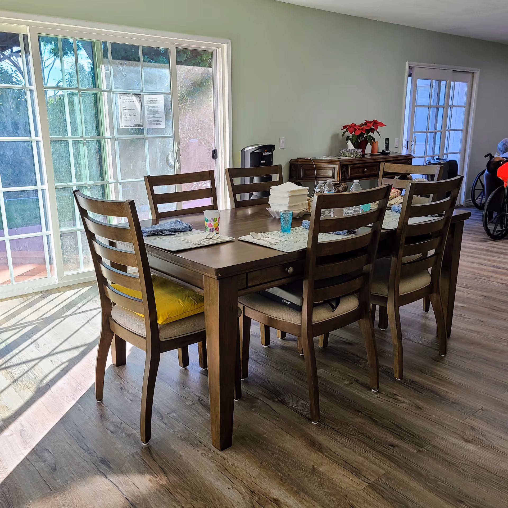 A dining area with a wooden table and six chairs, set with placemats, napkins, cups, and water bottles. The room has large sliding glass doors letting in natural light, a wooden sideboard with a red poinsettia plant, and a person in a wheelchair partially visible in the background near another door.
