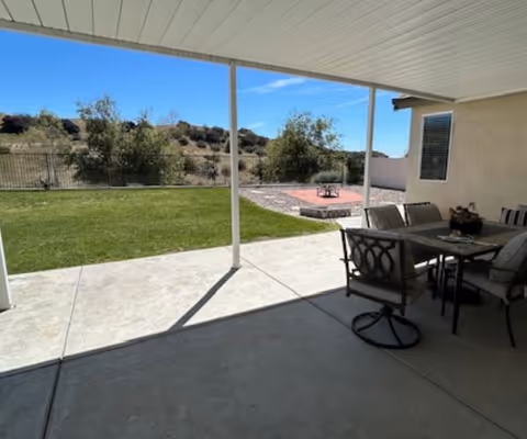 Covered patio with outdoor dining furniture overlooking a grassy backyard, a small fire pit area, and hills in the distance.