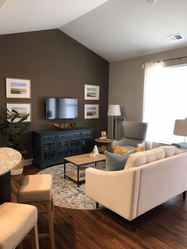 A cozy living room with a beige sofa adorned with colorful pillows, a gray armchair, a wooden coffee table on a patterned round rug, a dark green cabinet with a flat-screen TV mounted above it, four framed landscape pictures on the wall, a floor lamp, and a large window with sheer curtains letting in natural light. Two beige bar stools are partially visible in the foreground.