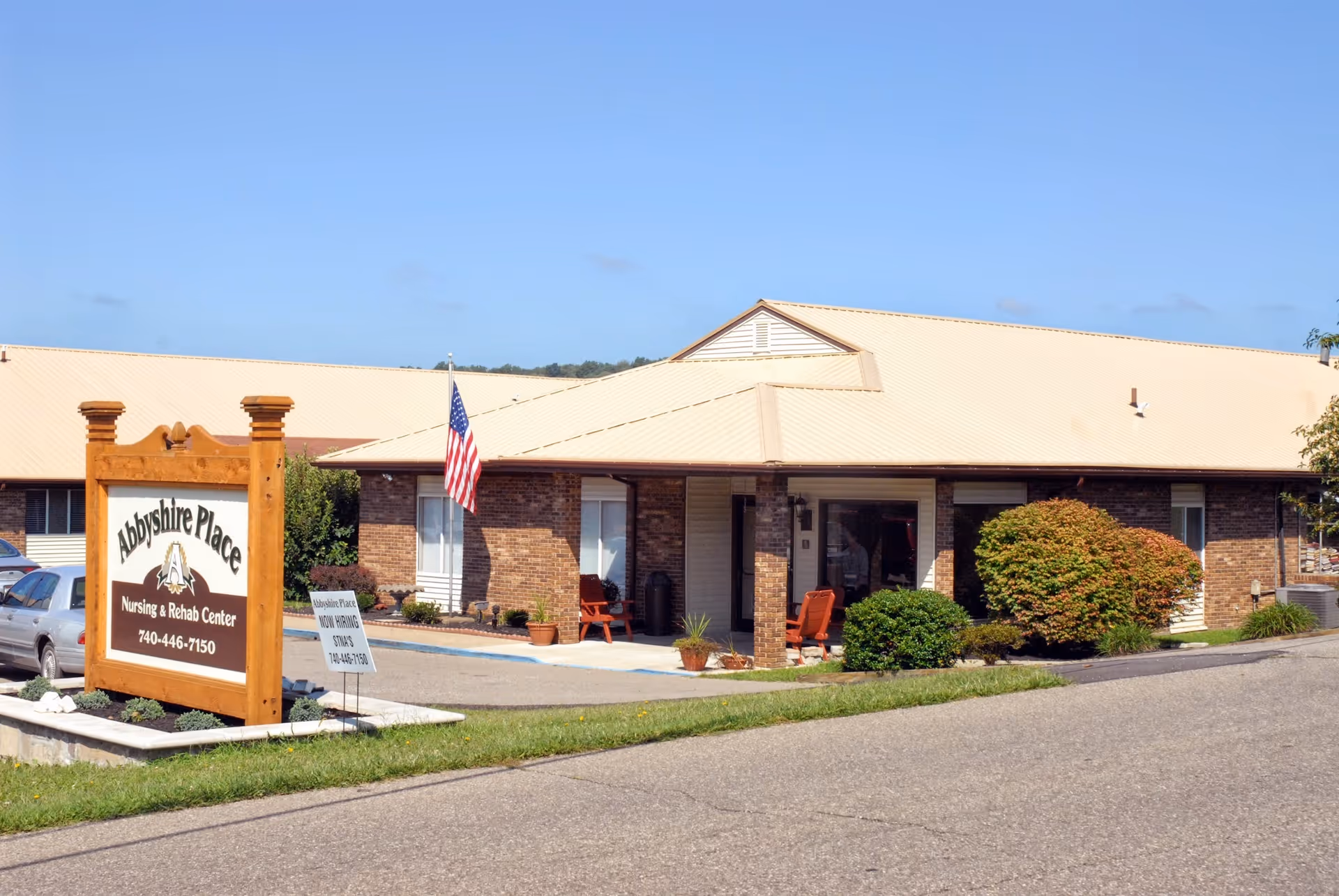 Exterior view of Abbyshire Place Nursing & Rehab Center, a single-story brick building with a beige roof. There is a wooden sign in front displaying the facility's name and phone number, an American flag on a pole, some bushes, and a paved driveway.