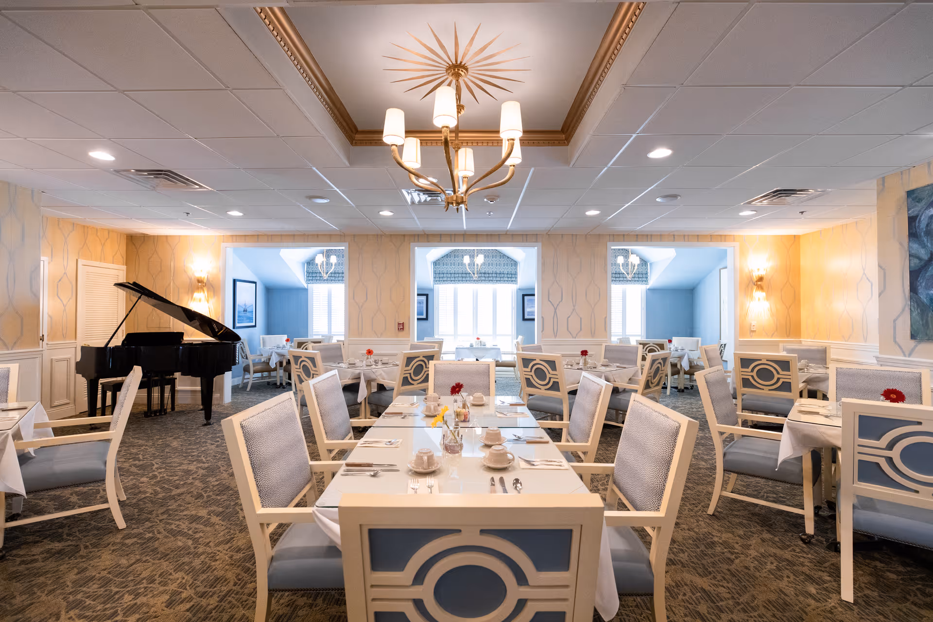 Spacious dining room with neatly set tables and upholstered chairs, a chandelier overhead, and a black grand piano in the corner.