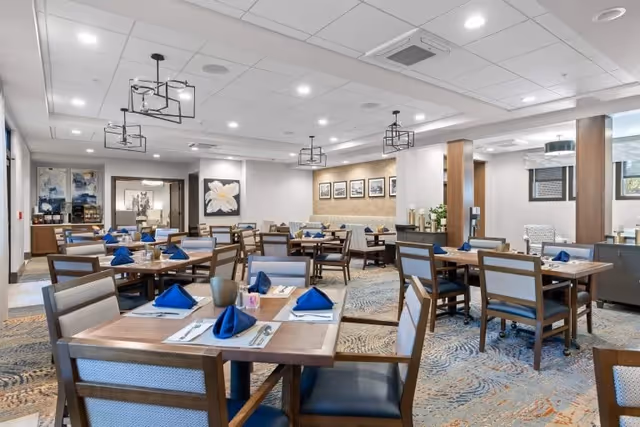 A spacious dining room with multiple wooden tables and chairs arranged neatly. Each table is set with blue folded napkins, placemats, and cutlery. The room features modern light fixtures hanging from the ceiling, framed artwork on the walls, and a patterned carpet. Large windows allow natural light to brighten the space.