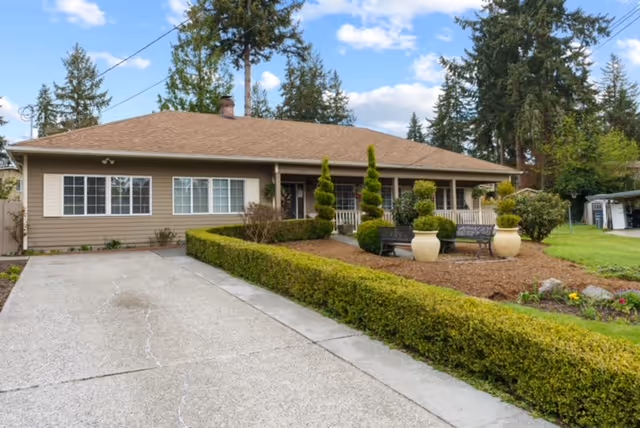 Single-story ranch-style house with a driveway, manicured hedges, and a front porch with seating in a landscaped yard.