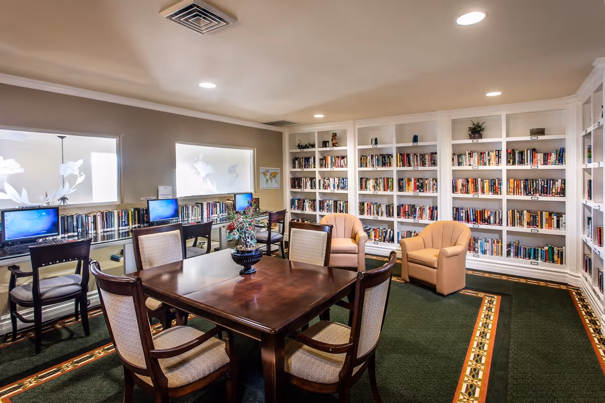 A cozy library room with built-in white bookshelves filled with books, two beige armchairs, a wooden table with four upholstered chairs, and three computer stations along one wall. The room has green carpet with a decorative border and recessed ceiling lights.