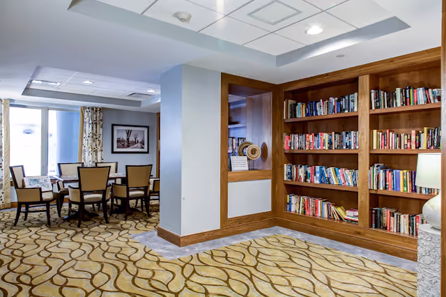 A well-lit senior living common area with built-in bookshelves and a dining table with chairs.