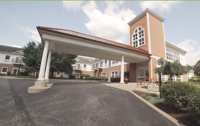 Exterior view of a senior living facility building with a covered entrance supported by white columns. The building is made of brick with white trim and has multiple windows. There is a sign near the entrance that reads 'The Suites Assisted Living'. The sky is partly cloudy.