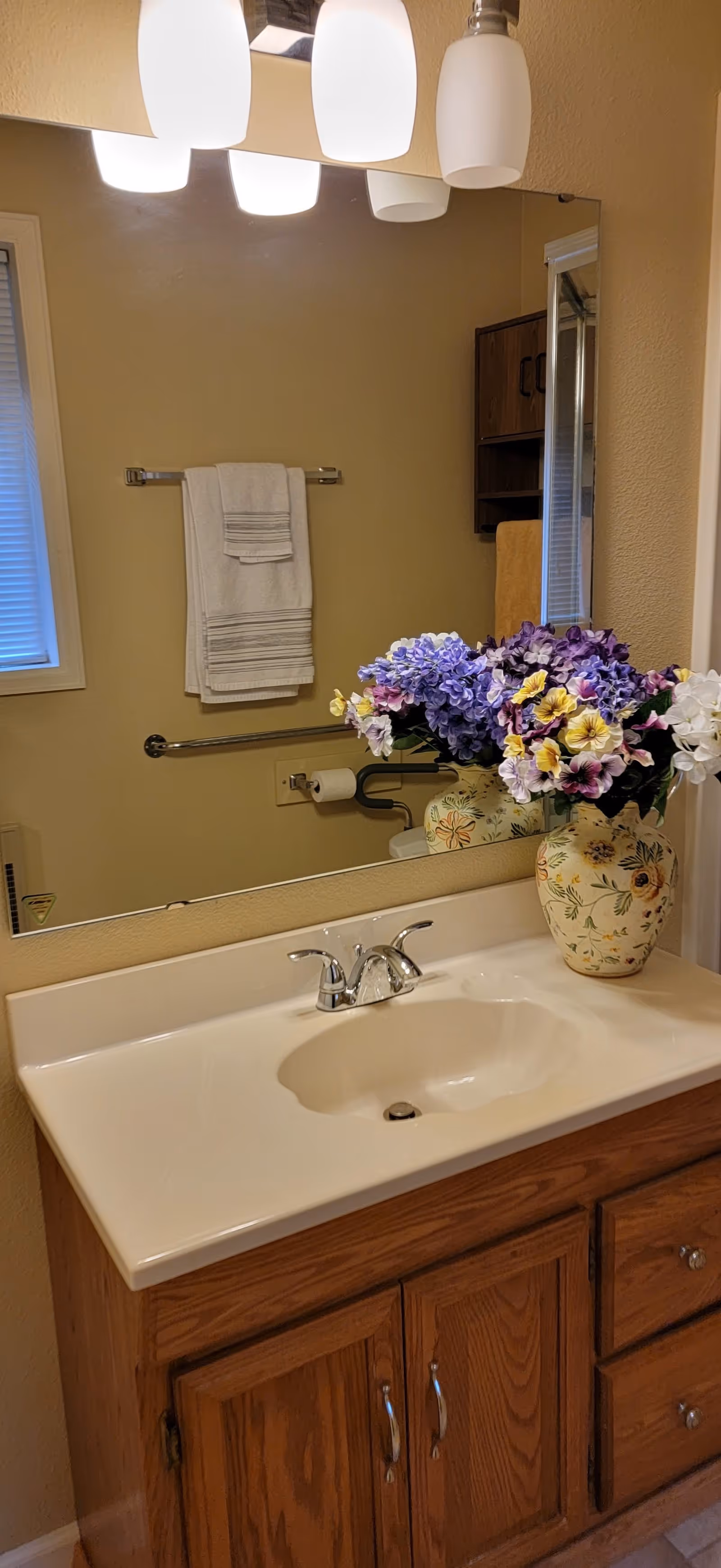Bathroom sink with a large mirror above it, a vase with purple and yellow flowers on the countertop, a towel rack with white towels, and a window with blinds partially visible.