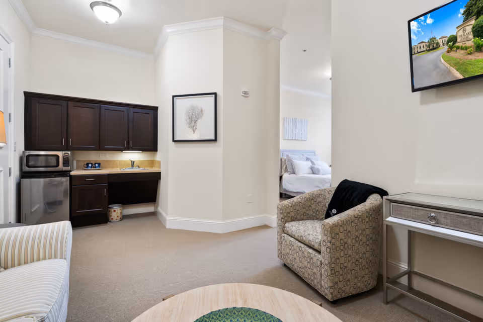 Interior view of a senior living facility suite showing a small kitchenette with dark wood cabinets, a microwave, and a mini fridge. There is a patterned armchair with a black blanket draped over it, a round wooden coffee table, and a striped armchair partially visible. A bedroom with a made bed is visible through an open doorway. A flat-screen TV mounted on the wall displays an image of the facility's exterior entrance. The walls are painted light beige and the carpet is light brown.