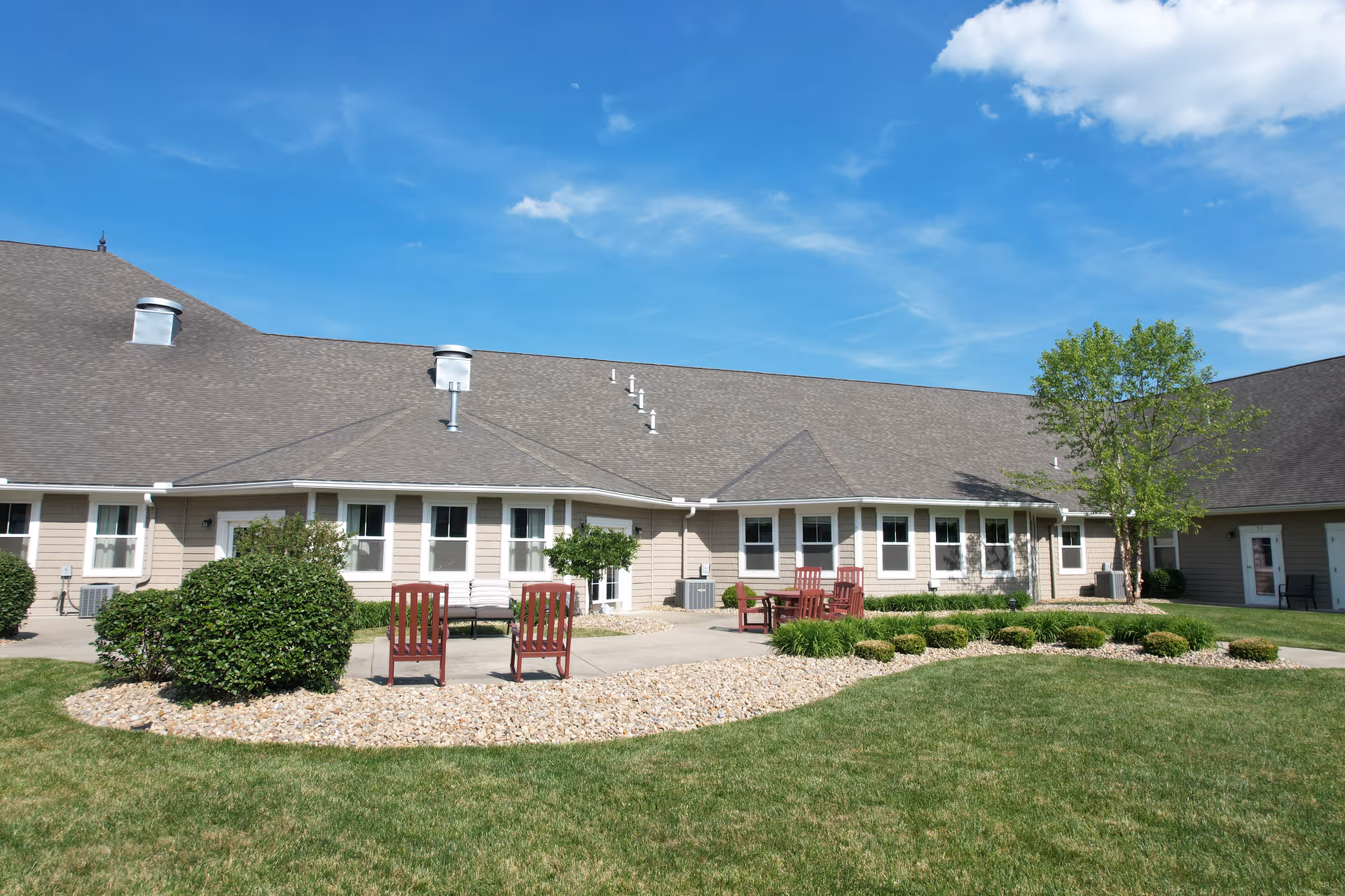 Outdoor view of a senior living facility with a beige building featuring multiple windows and a sloped roof. There is a well-maintained lawn, bushes, and small trees in front of the building. Several red wooden chairs and a bench are placed on a concrete patio area surrounded by decorative rocks. The sky is clear with a few clouds.