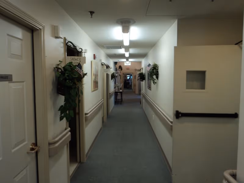 A long, narrow hallway in a senior living facility with beige walls and carpeted floor. The hallway is decorated with small plants and American flags mounted on the walls. Several doors line the hallway, one labeled 'PRIVATE'. Handrails run along both sides of the corridor, and fluorescent lights illuminate the space from the ceiling.