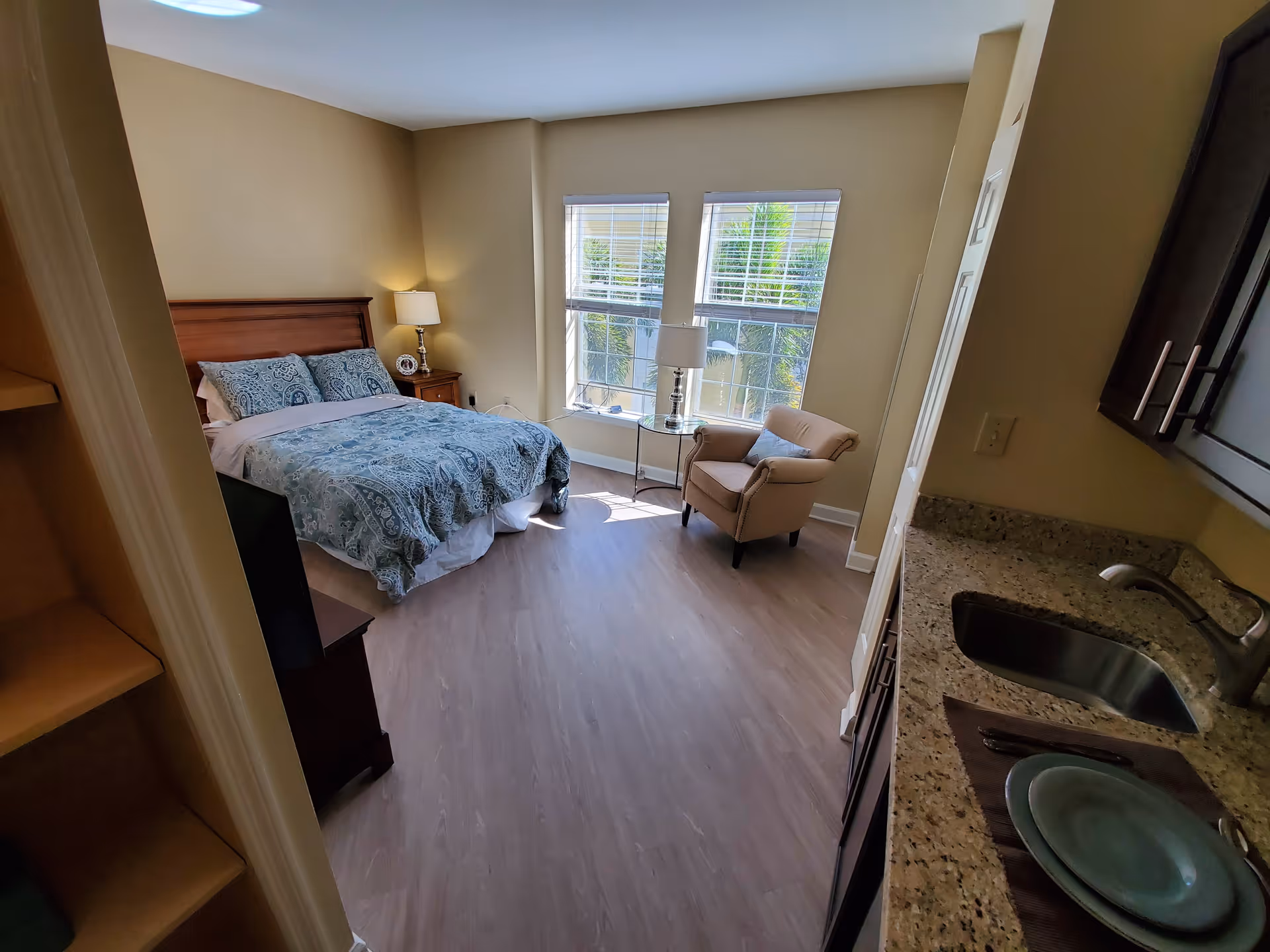 A bright bedroom with a bed covered in blue patterned bedding, a wooden nightstand with a lamp and clock, a beige armchair next to a small round glass table with a lamp, and two large windows with white blinds letting in natural light. To the right, there is a small kitchenette area with a granite countertop, sink, and dark cabinets.