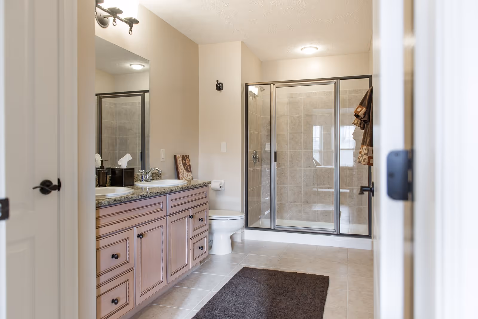 A clean and modern bathroom featuring a double sink vanity with granite countertop, a large mirror, a toilet, and a glass-enclosed tiled shower. The floor is tiled and there is a dark brown rug in front of the vanity.