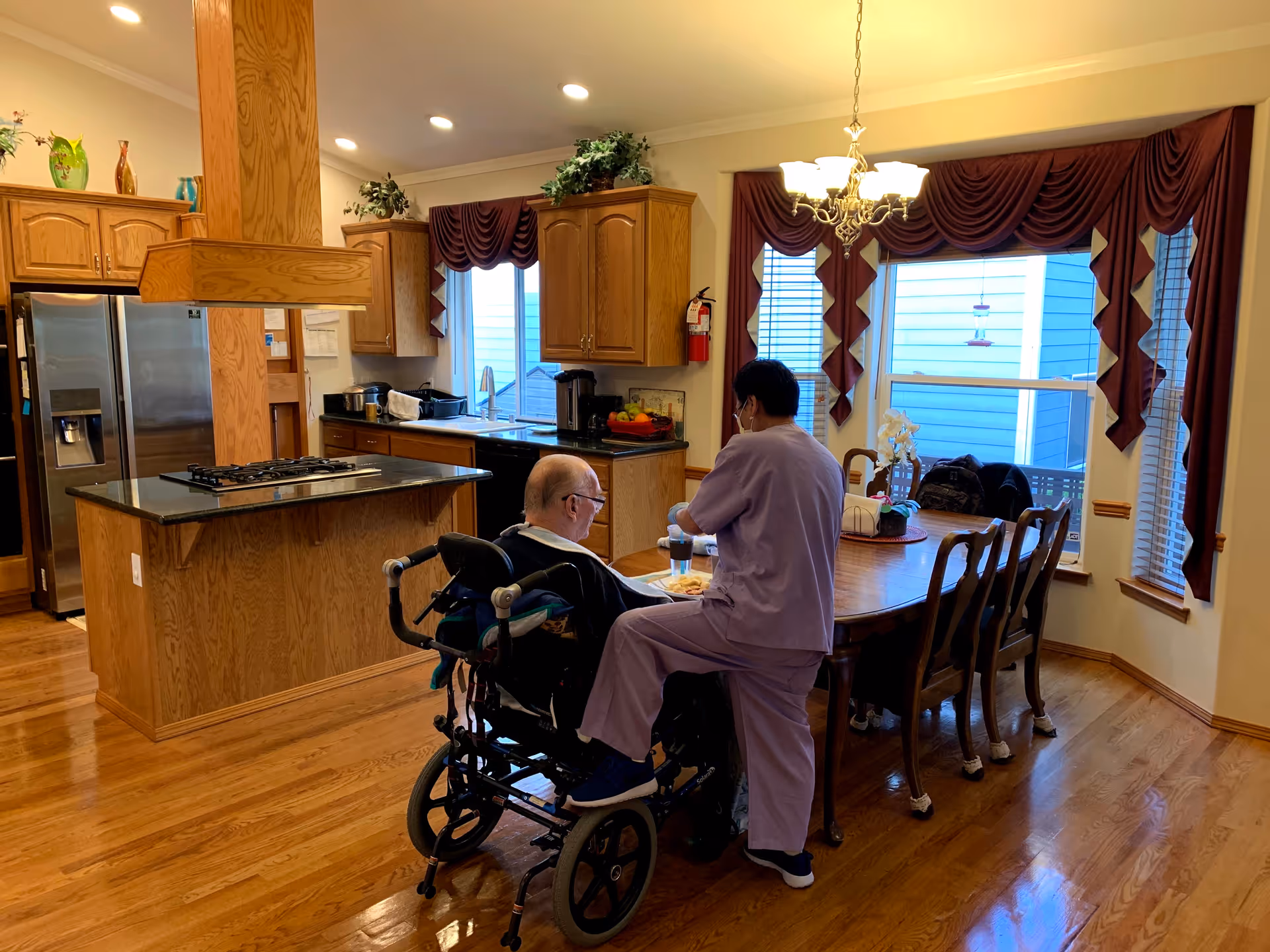 A caregiver assists an elderly man in a wheelchair at a dining table in a combined kitchen and dining area.