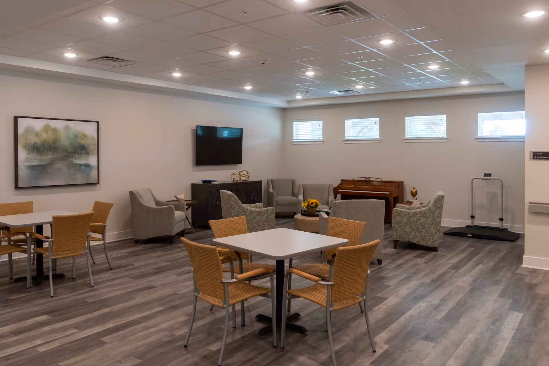 A spacious common area in a senior living facility featuring several tables with chairs, a seating area with armchairs, a piano, a wall-mounted TV, and a large painting on the wall. The room has wood-style flooring and multiple small windows near the ceiling allowing natural light.