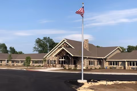 Front exterior of a single-story retirement facility with a stone facade, covered entrance, and an American flag on a flagpole.