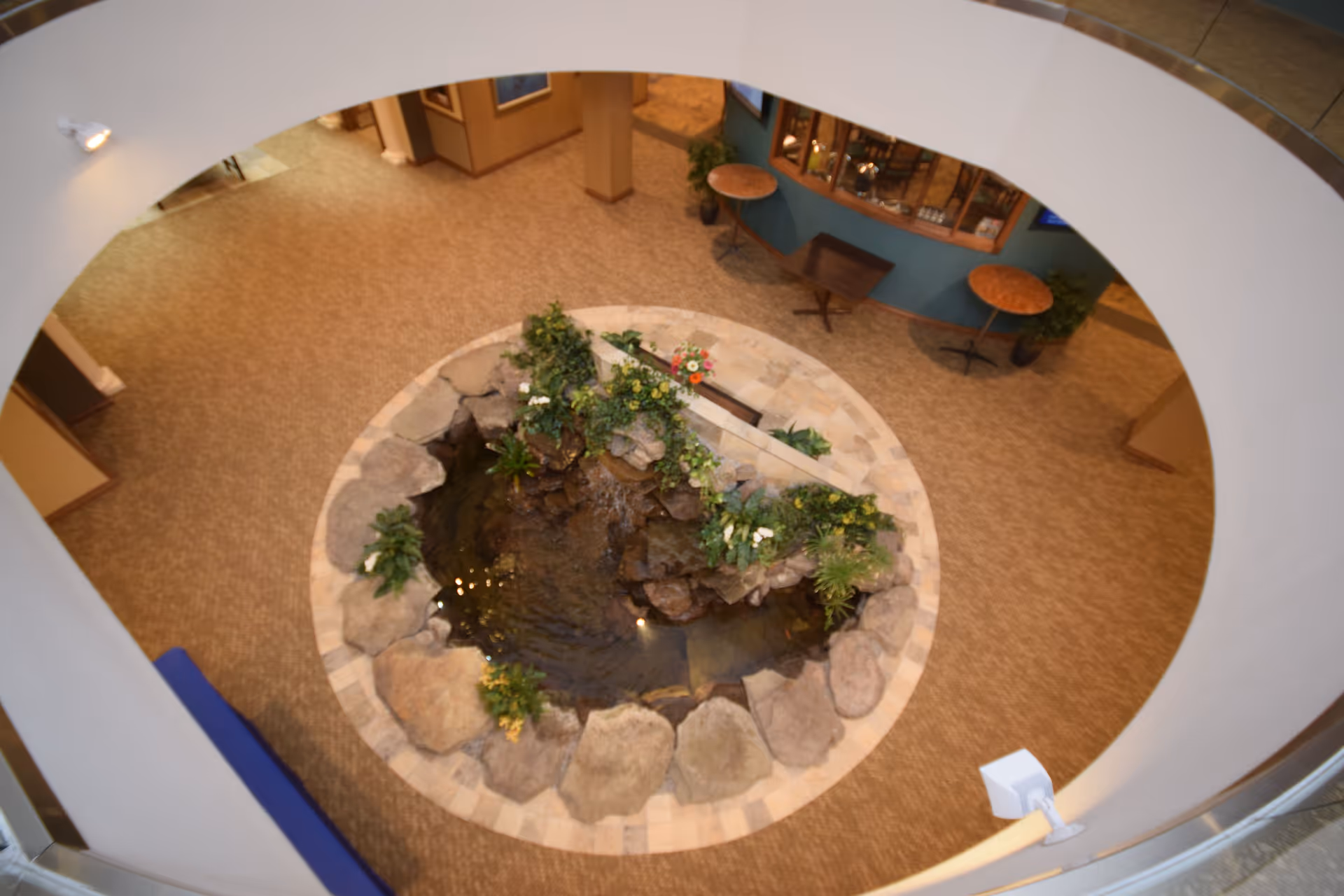 Top-down view of an indoor circular water feature with rocks and plants in the center of a carpeted area. Surrounding the water feature are small tables and chairs against a blue wall with a display cabinet. The image is taken from an upper floor looking down through a circular opening.