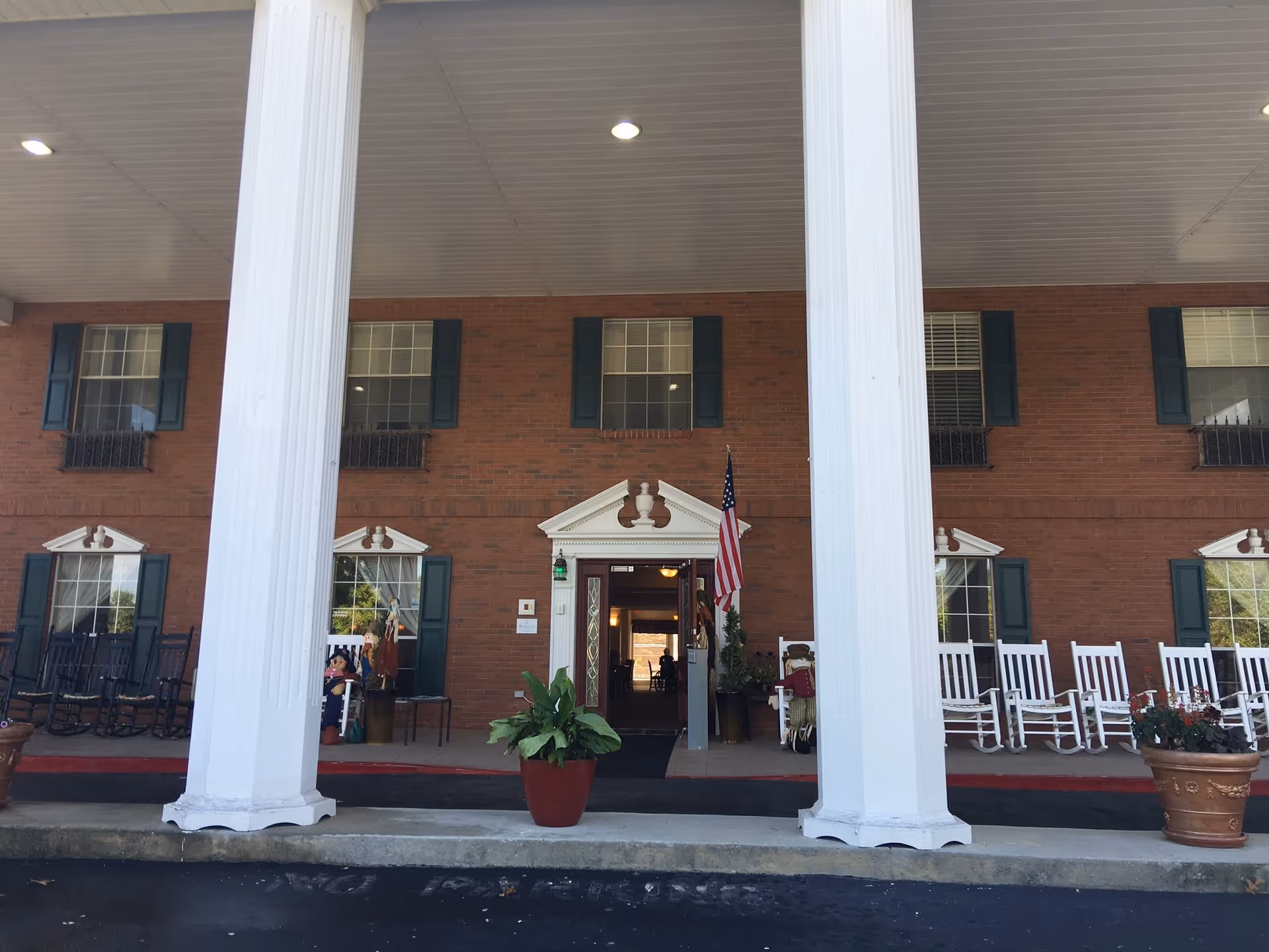 Front entrance of a brick building with large white columns, an American flag, potted plants, and white rocking chairs on the porch.