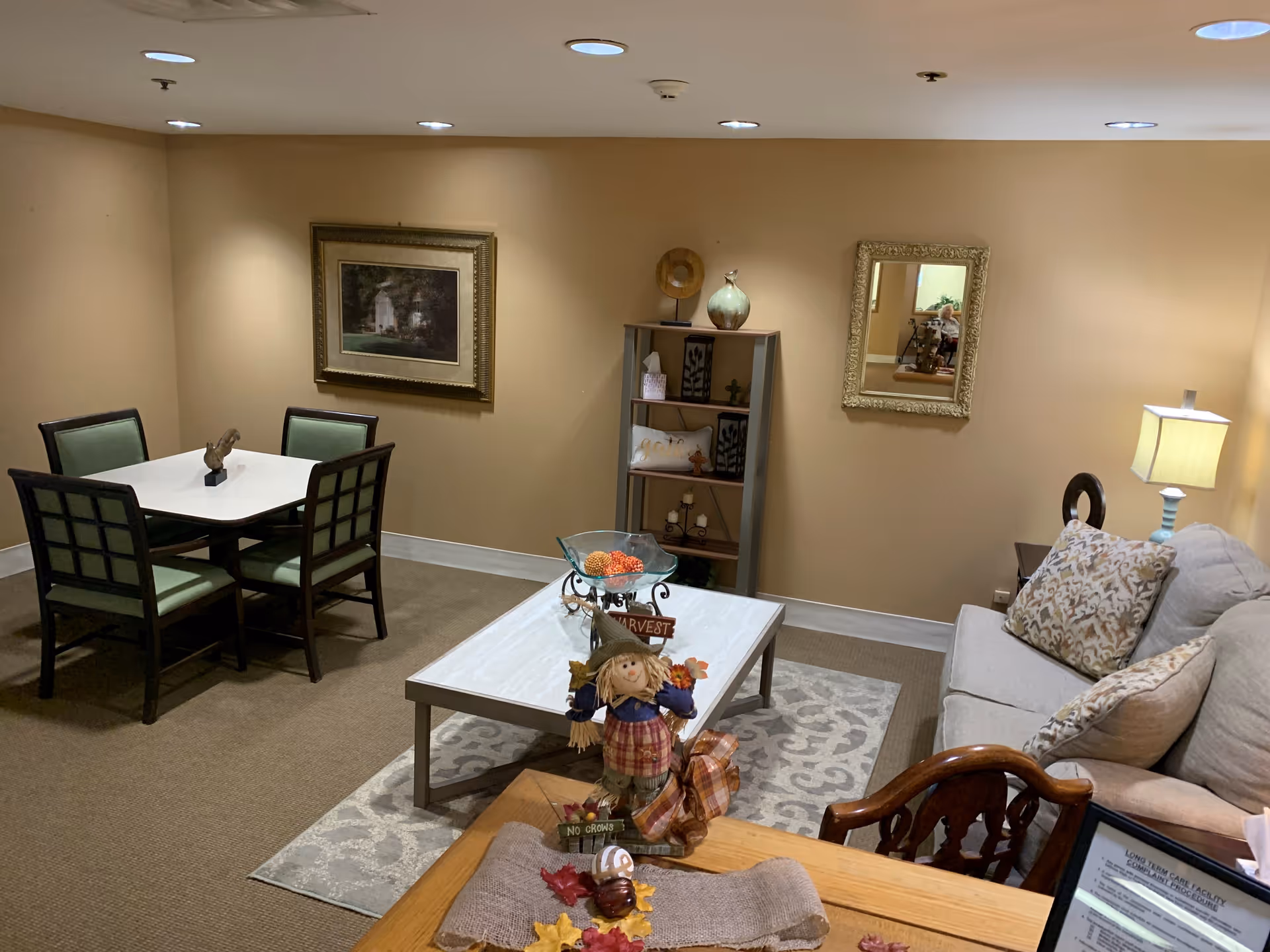 A cozy living room area with a beige couch adorned with patterned pillows, a wooden chair, a coffee table with decorative items including a scarecrow figure, and a small dining table with four green cushioned chairs. The walls are painted light brown and decorated with a framed picture and a mirror. A bookshelf with decorative items and a lamp on a side table are also visible.