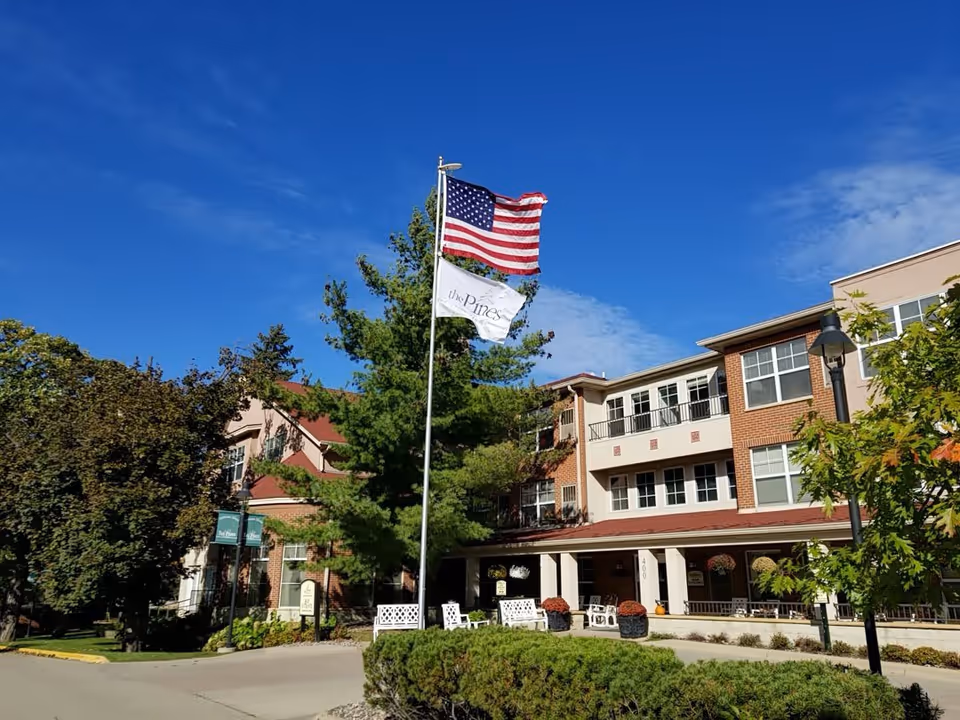 Exterior view of The Pines Senior and Assisted Living facility with a clear blue sky, an American flag and a white flag with the facility's name flying on a flagpole, surrounded by trees and bushes. The building is a multi-story brick and beige structure with balconies and a covered entrance area with white benches and potted plants.